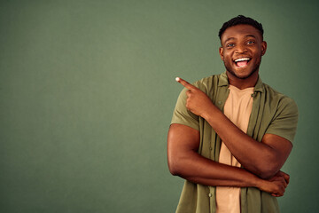 Advertising product. Positive young guy in casual khaki shirt pointing with finger on green background with copy space. Excited african man presenting promo offer or holiday discount in studio.