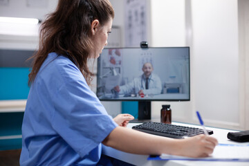 Nurse in videocall taking notes from doctor colleague in professional medical office. Woman writing...