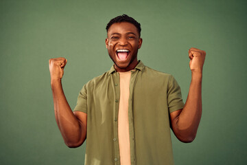 Celebration of victory. Emotional african american man in casual clothes making winner gesture with clenched fists while sincerely rejoicing big luck. Isolated over green studio background. - Powered by Adobe