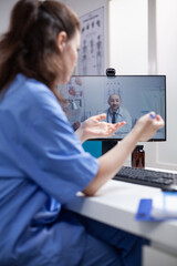 Nurse at desk in telemedicine videocall communicating with doctor specialist in professional medical office. Medic having online internet video conference with young colleague