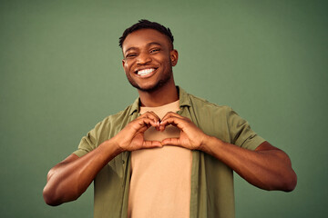 Expression of love. African american handsome guy smiling sincerely and making heart shape with hands over green background. Young male person in khaki shirt showing romantic feelings in studio. - obrazy, fototapety, plakaty
