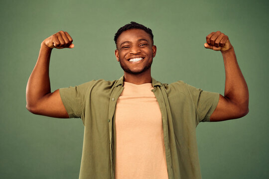 Result of regular workout. African american man wearing casual clothes smiling and showing biceps at camera over green studio background. Cheerful young guy satisfied with body and health condition.