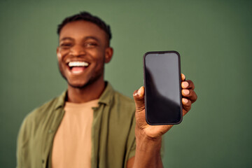 App advertisement. Blurred background of joyful african american guy standing over green background and showing modern smartphone with empty black screen. Focus on cell phone with space for text.