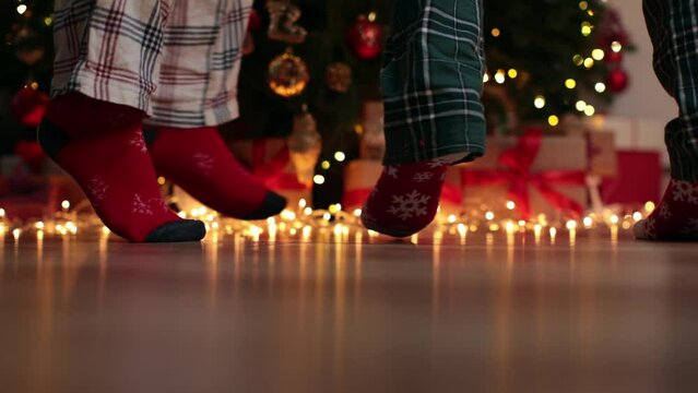 Christmas party celebration New Year indoors happy dancing legs and feet in funny Christmas socks and green red pajamas near decorated tree with blinking yellow magic lights on wooden floor concept