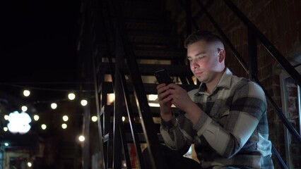 A young man sits on a fire escape and texts on a smartphone. A man on a narrow street late at night with a phone in his hand.