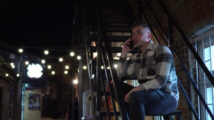 A young man sits on a fire escape and talks on the phone. A man on the stairs of a narrow street late at night. - obrazy, fototapety, plakaty
