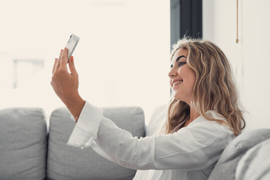 Selfie picture of happy beautiful young woman looking at camera with toothy smile. Pretty girl holding gadget with webcam, making video call, self home portrait. Communication concept.
