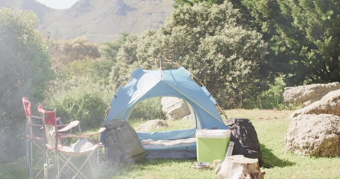 Smoking firepit, tent, chairs and camping equipment in sunny countryside, copy space, slow motion