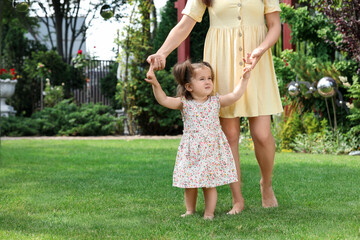 Mother supporting baby girl while she learning to walk on green grass in park