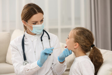 Doctor in medical mask examining girl`s oral cavity with tongue depressor indoors