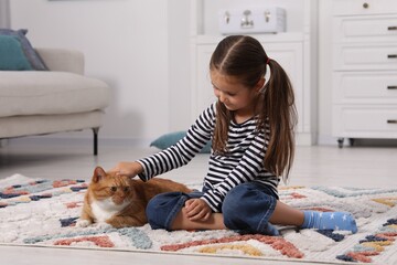 Little girl petting cute ginger cat on carpet at home