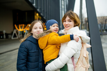 Two big sisters and their toddler brother having fun outdoors. Two young girls holding their sibling boy on winter day.