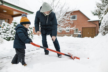 Adorable toddler boy helping his grandfather to shovel snow in a backyard on winter day. Cute child wearing warm clothes playing in a snow.