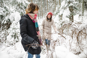 Cute teen girl and her mother having fun on a walk in snow covered pine forest on chilly winter day. Teenage child exploring nature.