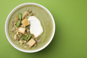 Delicious broccoli cream soup with croutons, sour cream and pumpkin seeds on green background, top view. Space for text