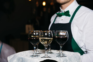 The waiter hold tray with several glasses of red and white wine. The employee wears a black apron, white shirt and green bowtie.