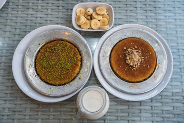 Two plates of Turkish kunefe topped with pistachio and walnut, with a glass of milk and banana.