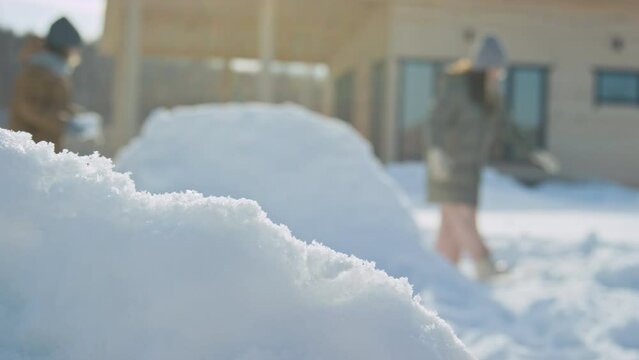 Close-up shot with lens flare of hands of unrecognizable child in winter jacket, woolly gloves and scarf making snow castle in back yard with friends on sunny day