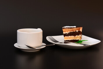 A piece of puff pastry cake with mint on a plate, a fork and a cup of hot coffee on a black background.