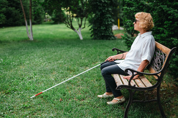 An elderly blind woman sits on a bench in the park.
