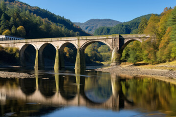 A bridge over a river in a mountainous area. The bridge is made of stone and has multiple arches, and its reflection can be seen in the calm, clear water below