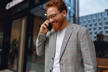 Bearded male freelancer in eyeglasses is talking phone standing in modern coworking space