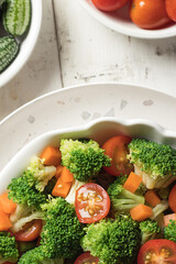 White wooden table with a vegan salad with pieces of broccoli, tomato, pickles and carrot.