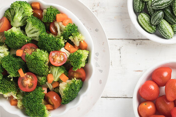 White wooden table with a vegan salad with pieces of broccoli, tomato, pickles and carrot.