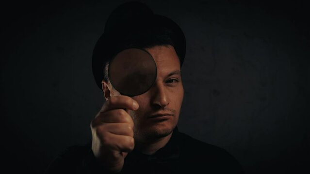 brunette man in a black hat with a magnified glass on a dark background close-up cinematic 4K 24 fps