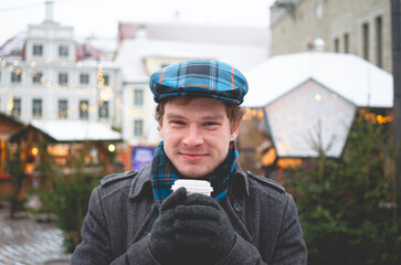 A handsome young man in a grey coat with a Scottish tweed cap and tartan scarf holding cocoa cup...