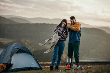 Fototapeta premium Diverse couple, putting on warm clothes, enjoy camping by a lake with a stove