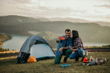 A couple using a phone, relaxes around a campfire on a hilltop, overlooking a serene lake