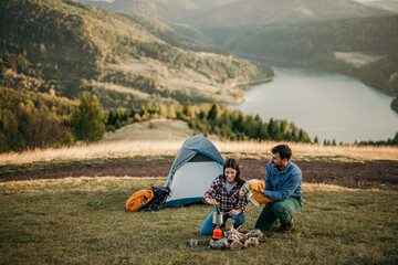A couple's campsite on a hill offers a view of a peaceful lake as they cook dinner and dress up.