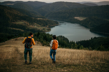 Fototapeta na wymiar Two friends walking uphill with backpacks, catching sight of a serene lake
