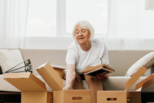elderly woman sits on a sofa at home with boxes. collecting things with memories albums with photos and photo frames moving to a new place cleaning things and a happy smile. Lifestyle retirement.