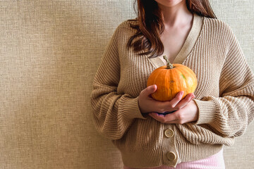 An unrecognizable brunette girl in a beige sweater holds a bright orange pumpkin in her hands on a beige background