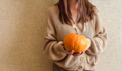 An unrecognizable brunette girl in a beige sweater holds a bright orange pumpkin in her hands on a beige background