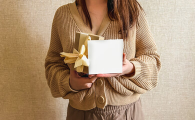 An unrecognizable brunette girl in a beige sweater holds a white square mockup and a gift in her hands. An empty sign with a gift in the hands of a girl