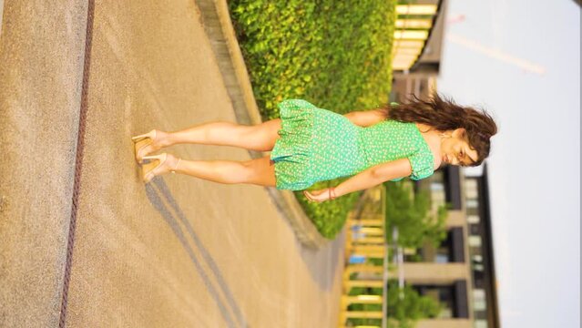perfect brunette model in her 20s walking through modern park with a wide footpath wearing heels and cute short dress posing looking over her shoulder into your eyes with interest slow motion vertical