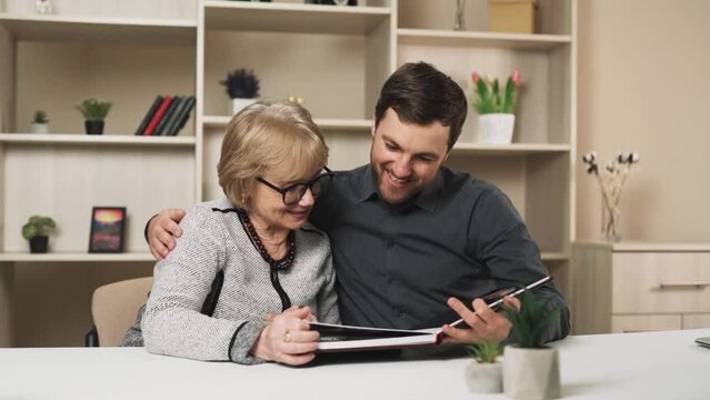 young man and an elderly woman in glasses embrace and cheerfully review memories in a printed photo album while sitting at the table