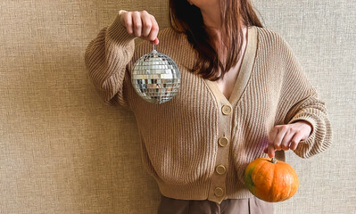 An unrecognizable brunette girl in a beige sweater holds a silver disco ball and an orange pumpkin in her hands. Halloween or Thanksgiving House Party