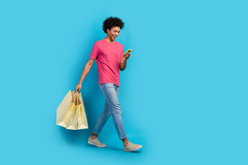 Full size photo of attractive young man walk hold device shopping bags wear trendy pink clothes isolated on blue color background