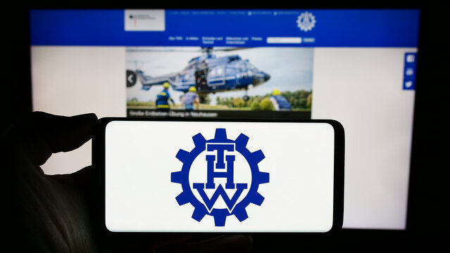 Stuttgart, Germany - 10-08-2023: Person holding mobile phone with logo of organization Bundesanstalt Technisches Hilfswerk (THW) in front of web page. Focus on phone display.
