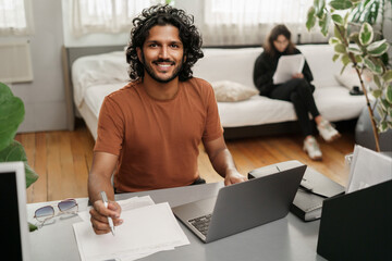Joyful student on his laptop, with a woman handling papers behind, all in a cozy ambiance