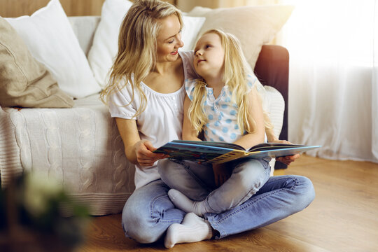 Happy family. Blonde young mother reading a book to her cute daughter while sitting at wooden floor in sunny room. Motherhood concept