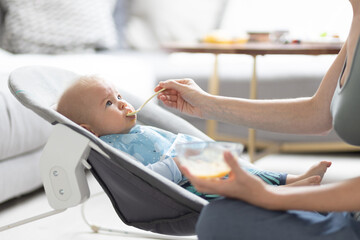 Obraz na płótnie Canvas Mother spoon feeding her baby boy child in baby chair with fruit puree. Baby solid food introduction concept