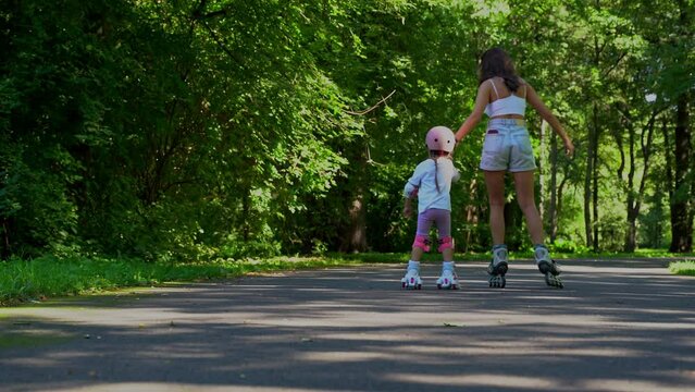 Mom teach little daughter girl inline skating with roller in public park in summer sunny day. Happy family time together, Leisure sport activity.