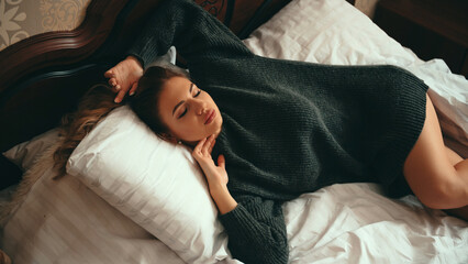 Beautiful young woman with bare legs, wearing green sweater, relaxed lies on a wooden bed with eyes closed and smiling. Resting and comfort concept. - obrazy, fototapety, plakaty