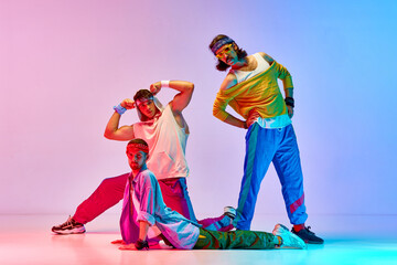 Funny young men in stylish colorful vintage sportswear doing aerobics exercises against gradient pink blue background in neon light. Concept of sportive and active lifestyle, humor, retro style. Ad
