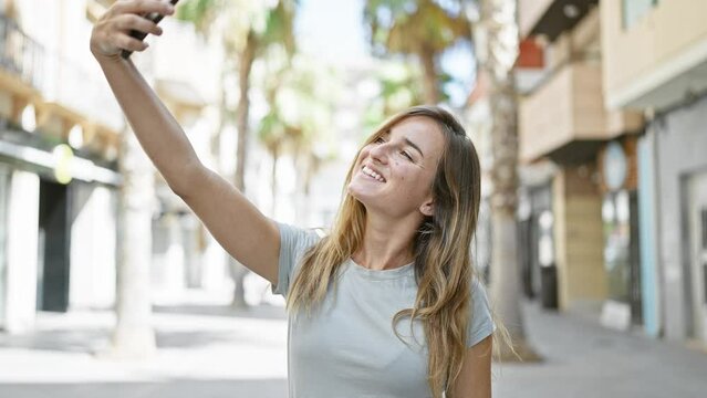 Joyful young blonde woman confidently making a fun selfie with her smartphone on the city street, basking in sunlight while enjoying her urban lifestyle.